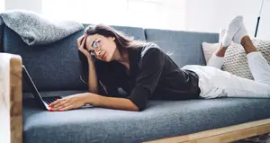 A woman is lying on a couch with a laptop on her lap. She has a gray blanket on the back of the couch, and a white pillow is placed beside her. The woman is wearing glasses and a black long-sleeve shirt. She is holding a cell phone in her hand and seems to be talking to someone. She is wearing white socks and a white bracelet.