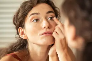 A woman is looking at her reflection in a mirror while applying an eye contact lens.