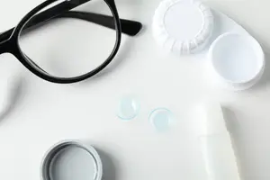 Black glasses, two contact lenses, and a bottle of contact lens solution on a white table