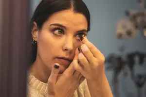 A woman wearing a white sweater and earrings is applying a contact lens in front of a mirror.