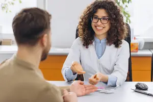 A woman in a white lab coat sitting at a desk talking to a man in a brown shirt.