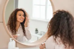 A woman with curly hair is smiling at her reflection in a mirror while holding a toothbrush.