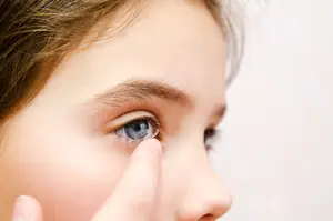 A girl with brown hair is trying to put on a contact lens in her right eye while looking at something in front of her.