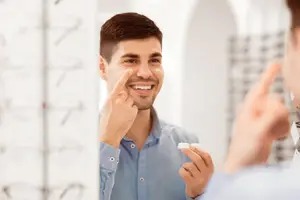 A man smiles and touches his face while holding a contact lens in his hand