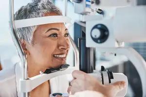 A woman with gray hair is smiling and looking at the camera while wearing a white headband and a white shirt, she is also wearing an eyeglass and seems to be checking her eye with an eyeglass machine, a person is standing on the right side, probably adjusting the machine, and there is a blurry background behind them.