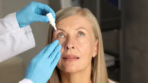 A woman is receiving eye drops from a doctor in a white coat, with blue gloves, and is looking at the doctor. The woman is wearing earrings and has blonde hair, and is looking at the doctor, who is holding a bottle of eye drops.