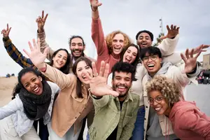 A group of people are smiling while posing for a photo on the beach, with one person sitting on another's shoulders, and another person standing in front of them, with a tower in the background.