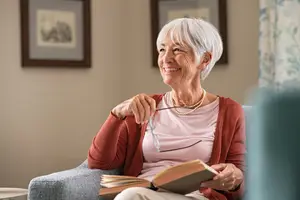 An elderly woman sitting on a chair, smiling and reading a book.