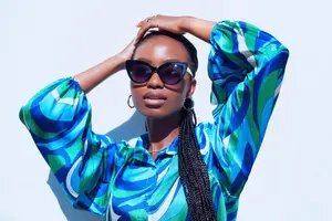 African woman posing for a photo wearing a blue and green patterned shirt, sunglasses, and earrings