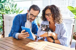 A man and a woman sitting on a chair in front of a table, looking at a tablet with a dog on the woman's lap.