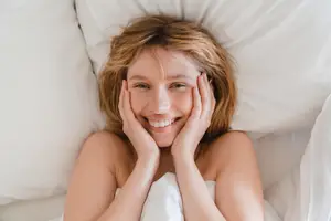A woman with blonde hair lying on a bed with a white blanket, smiling and posing for a photo.