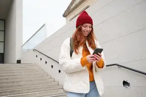 A woman in a white jacket and red beanie smiles and looks at her phone while standing near stairs in a building.