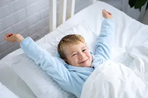 A little boy is lying in bed and smiling with his arms raised