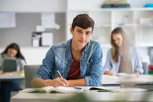 A student in a denim jacket is seated at a desk in a classroom, surrounded by other students, and they are all engaged in studying with books and notebooks open in front of them.