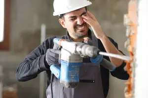 Man in a white helmet holding a drill while holding his face with his hand