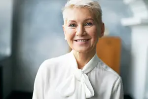An adult woman with short blonde hair wearing a white blouse with a bow tie is smiling and standing in front of a wooden cabinet in an indoor area.