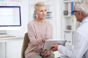 An elderly woman sits on a chair while looking at a man in a white coat who holds a tablet in a hospital room