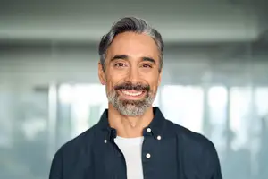 A smiling man with gray hair and a beard wearing a navy shirt is standing in a room with a glass wall.