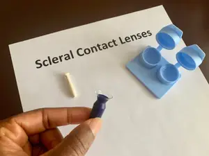 A person is holding a blue contact lens in their hand while a blue lens case is on a white paper with the text Scleral Contact Lenses.