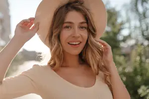 A woman with blonde hair is smiling and posing for a photo while holding her hat.