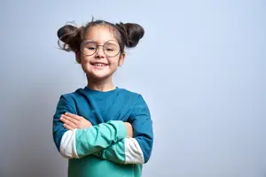 A young girl with glasses and a blue shirt is smiling and posing for a photo with her arms crossed.