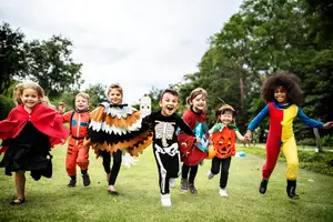 A group of children in Halloween costumes running on a grassy field with trees in the background.