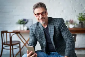 A smiling man with glasses is sitting in a chair and looking at his phone