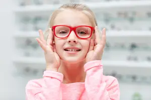 A young girl with blonde hair and red glasses is smiling and posing for a photo in a room with shelves in the background.