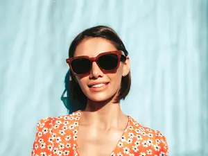 A smiling woman with short hair and glasses with a brown frame and black lenses, wearing an orange floral blouse, standing in front of a light blue wall.