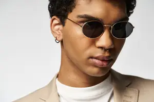 A young man with a brown coat, a white shirt, and sunglasses with a black lens and a brown frame. He is posing for a picture with his lips slightly open. He has a ring earring and a nose ring.