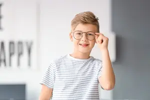 A boy in a striped white shirt adjusting his glasses in a room with a wall that says Happy