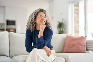 A woman is sitting on a couch with her hand on her chin and looking to the right. She is wearing a blue long-sleeve shirt, glasses, and a ring. Behind her is a window with a view of a plant and a brick wall. The room is well-lit, and there is a microwave and a cabinet in the background.