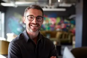 A smiling man in a black shirt stands in an office with colorful murals and furniture.