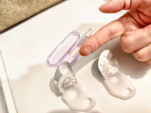 A hand is touching a clear plastic object, possibly a contact lens, on a white surface.