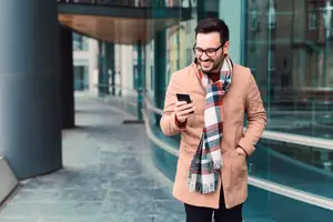 Man in a tan coat and scarf standing on a sidewalk looking at his phone.