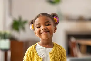 A young girl with a yellow cardigan and pink and yellow flower clips in her hair smiles in a living room