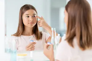 Woman in a bathroom applying eye drops in front of a mirror