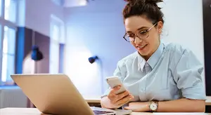 A woman wearing glasses and a blue shirt is smiling and looking at her phone while sitting in front of a laptop on a desk.