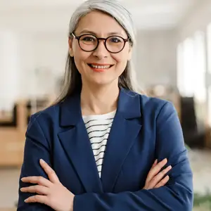 A woman with gray hair wearing glasses and a blue blazer is smiling and posing for a photo in an office.
