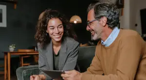 An adult man and woman are sitting on a couch, smiling, and looking at a tablet, with a wooden table, a potted plant, a lamp, and a black wall in the background.