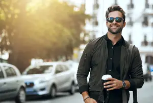 A man wearing sunglasses and a jacket is walking on a road, holding a cup in his left hand and smiling.