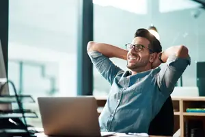 A man with glasses and a denim shirt relaxing with his hands behind his head in front of a laptop
