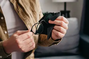 A man wearing a brown jacket is cleaning his black-framed glasses with a green cloth.