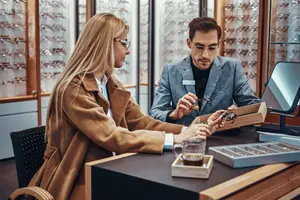 A man and woman are looking at glasses in a store, with a man holding a box of glasses and the woman holding a pair of glasses.