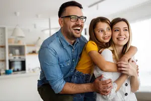 Three people in a living room with a smiling man holding a girl and a woman with blonde hair and a bracelet standing in front of a white shelf with a lamp