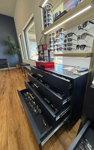 A black cabinet with four drawers filled with glasses and a glass top with magazines and books is placed on a wooden floor near the window. There is a wooden shelf with many pairs of glasses on top of the cabinet. A potted plant and a black bench are on the left side, and a red box is on top of the cabinet. On the right side, there is another cabinet with glasses.