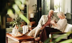 An elderly couple sitting on a couch and looking at each other while holding cups of coffee