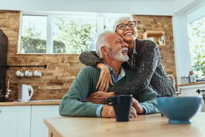 An elderly couple sitting in the kitchen, the man wearing a green sweater and the woman wearing a gray sweater, they are both smiling and the woman is hugging the man from behind, there is a table in front of them with a bowl, a cup, and a kettle on it