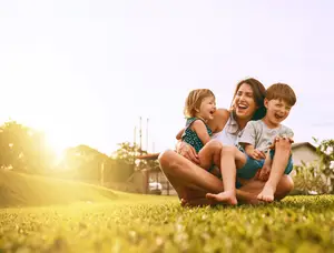 A mother and two kids sitting on a lawn, smiling and laughing at the camera, with a house and trees in the background