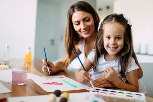 A woman and a young girl sitting at a table, painting together and smiling.